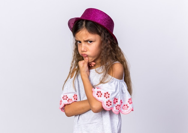 Foto grátis menina caucasiana irritada com chapéu de festa roxo segurando o queixo isolado na parede branca com espaço de cópia