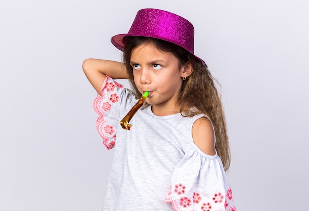 Foto grátis menina caucasiana irritada com chapéu de festa roxo, soprando apito de festa e olhando para cima, isolada na parede branca com espaço de cópia