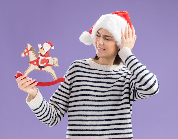 Menina caucasiana irritada com chapéu de papai noel coloca a mão na cabeça, segurando e olhando para o papai noel em decoração de cavalo de balanço isolada na parede roxa com espaço de cópia