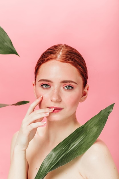 Menina caucasiana nua com folhas verdes, olhando para a câmera foto de estúdio de mulher ruiva espetacular com planta isolada em fundo rosa