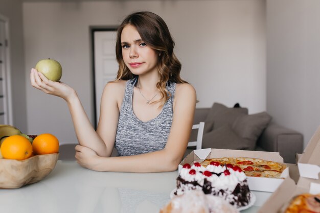 Menina caucasiana pensativa, pensando em sua dieta e segurando a maçã. Foto interna de mulher séria e encaracolada, posando à mesa com comida saudável e pizza.