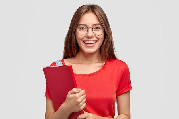 Menina caucasiana positiva com sorriso encantador, usa uma camiseta vermelha, segura um livro, modelos contra uma parede branca, tem disposição para estudar, usa óculos ópticos para uma boa visão. Juventude, conceito de aprendizagem