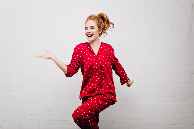 Menina caucasiana positiva magro em roupa de noite vermelha na moda dançando na parede de tijolos. Foto interna da bela jovem branca de pijama se divertindo em casa.