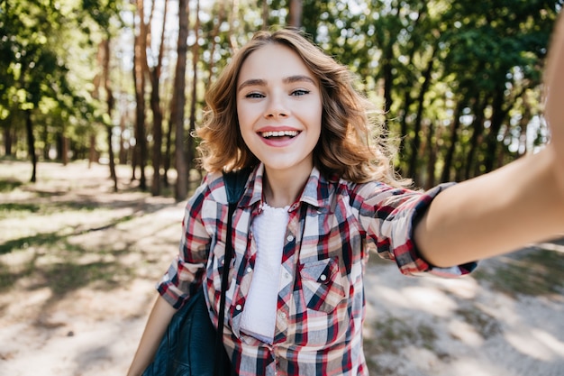 Menina caucasiana refinada em camisa quadriculada, caminhando na floresta. Retrato ao ar livre da senhora encaracolada rindo fazendo selfie em dia ensolarado.