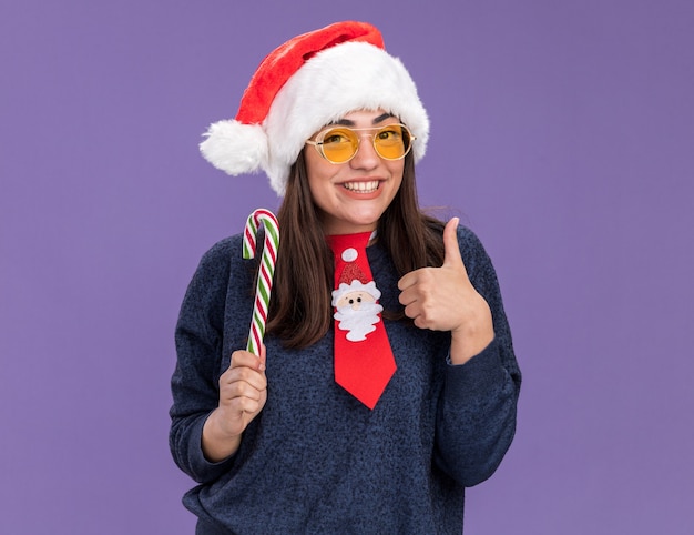 Menina caucasiana sorridente em óculos de sol com chapéu de Papai Noel e gravata de Papai Noel segurando o bastão de doces e polegares para cima, isolado na parede roxa com espaço de cópia