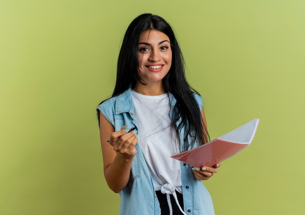 Foto grátis menina caucasiana sorridente segurando caneta e caderno isolados em um fundo verde oliva com espaço de cópia