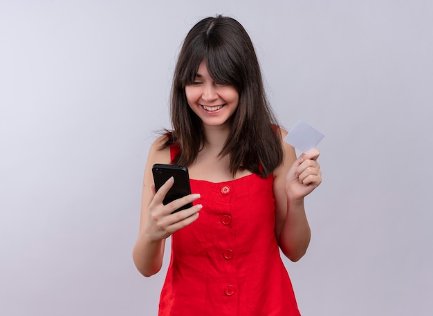 Menina caucasiana sorridente segurando um telefone e um cartão, olhando para o telefone, em um fundo branco isolado com espaço de cópia