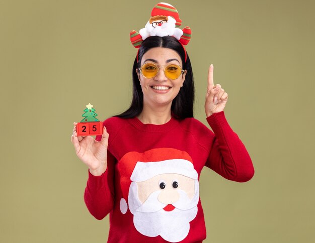 Menina caucasiana sorridente, vestindo uma bandana de Papai Noel e um suéter com óculos segurando um brinquedo de árvore de Natal com data olhando para a câmera apontando para cima isolado no fundo verde oliva