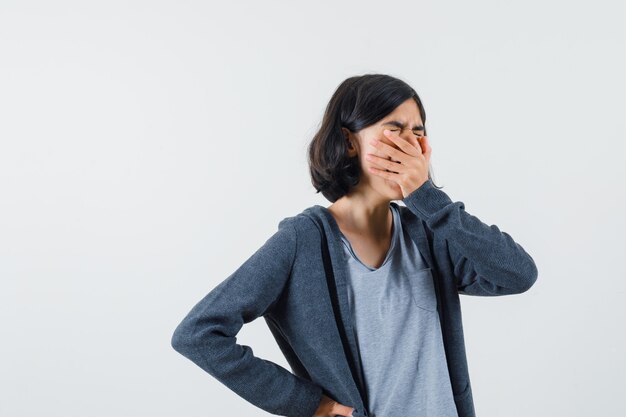 Menina cobrindo a boca com a mão, colocando a outra mão na cintura enquanto boceja com uma camiseta cinza claro e um capuz cinza escuro com zíper na frente e parecendo sonolenta