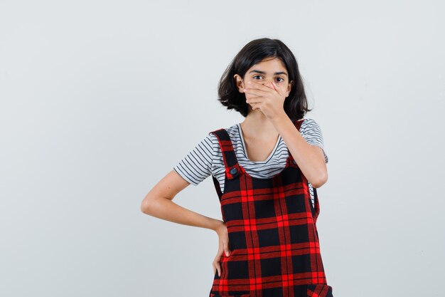 Menina cobrindo a boca com a mão na camiseta, macacão e parecendo preocupada,