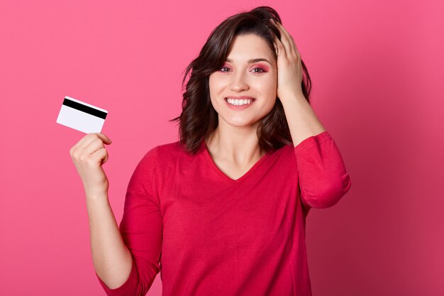 Menina com aparência agradável, segurando o cartão de crédito nas mãos, ganha dinheiro na loteria, mulher vestida de vermelho parece feliz, paga por compras com cartão, mantendo a palma da mão no rebanho.