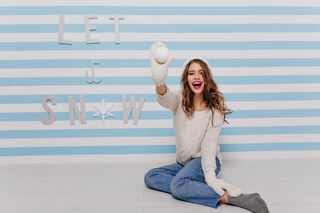 Menina com cabelo comprido e encaracolado e batom brilhante posando de brincadeira, jogando uma bola de neve em direção a