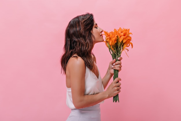 Foto grátis menina com cabelo ondulado gosta do perfume de lindas flores de laranja. mulher de cabelos escuros em roupa leve da moda cheira um grande buquê.