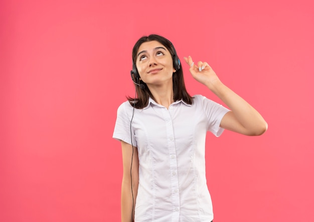Menina com camisa branca e fones de ouvido, fazendo um desejo desejável cruzando os dedos feliz e positivo em pé sobre a parede rosa