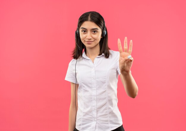 Menina com camisa branca e fones de ouvido, mostrando e apontando com os dedos para cima, número três, sorrindo em pé sobre a parede rosa