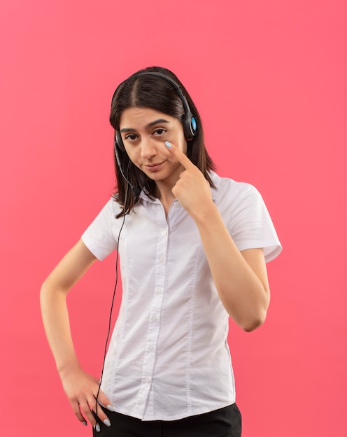 Foto grátis menina com camisa branca e fones de ouvido, olhando para a frente apontando para o olho, vendo você gesticular em pé sobre a parede rosa