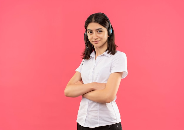 Menina com camisa branca e fones de ouvido, olhando para a frente com uma expressão confiante com os braços cruzados em pé sobre a parede rosa