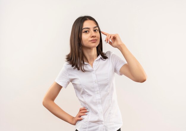 Menina com camisa branca olhando para a frente apontando para a têmpora e se concentrando em uma tarefa em pé sobre uma parede branca