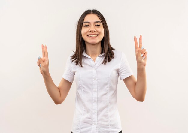 Menina com camisa branca sorrindo alegremente mostrando a vitória e cantando com as duas mãos em pé sobre a parede branca