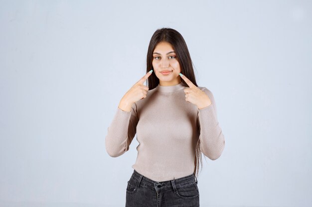 Menina com camisa cinza, apontando para a boca dela.