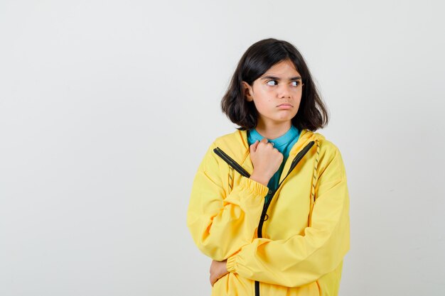 Menina com camisa, jaqueta, olhando para longe, mantendo a mão sobre o peito e olhando pensativa, vista frontal.