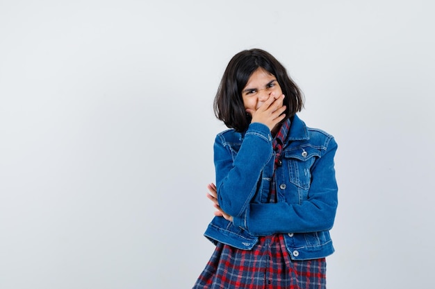 Menina com camisa, jaqueta, segurando a mão na boca e parecendo feliz, vista frontal.