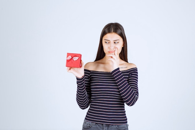 Menina com camisa listrada, segurando uma caixa de presente vermelha, parece confusa e duvidosa.