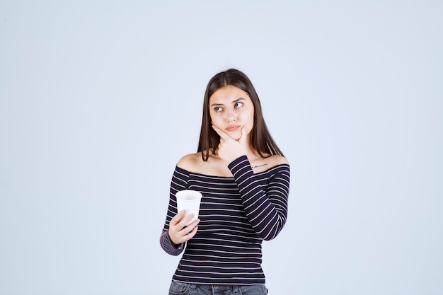 Menina com camisa listrada, segurando uma xícara de café de plástico e pensando.
