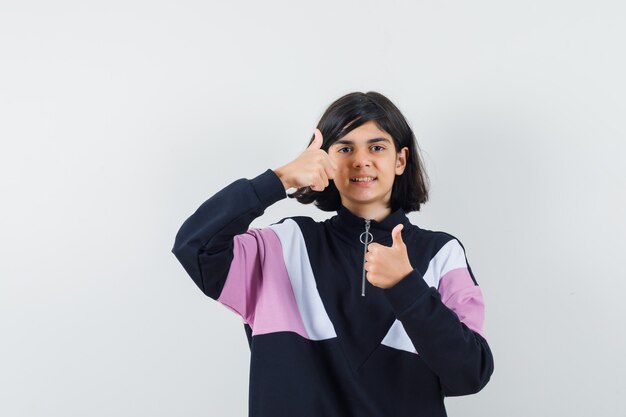 Menina com camisa, mostrando os polegares duplos e parecendo confiante, vista frontal.