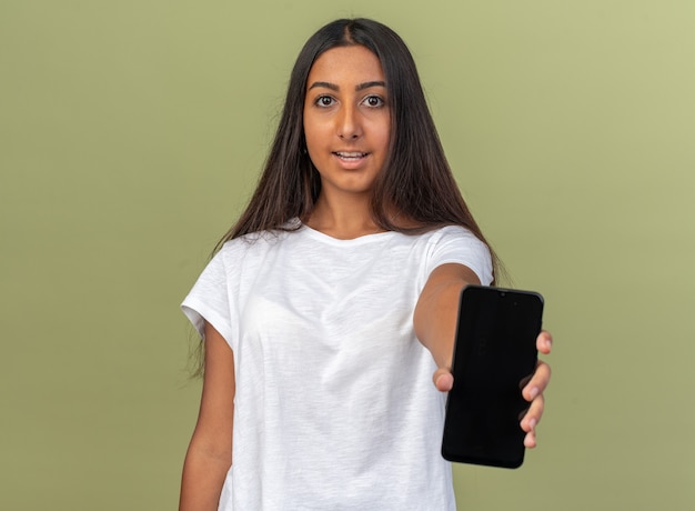 Menina com camiseta branca mostrando smartphone olhando para a câmera sorrindo confiante em pé sobre o verde