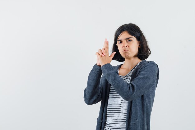 Menina com camiseta e jaqueta fazendo sinal de pistola com o dedo e parecendo rancorosa