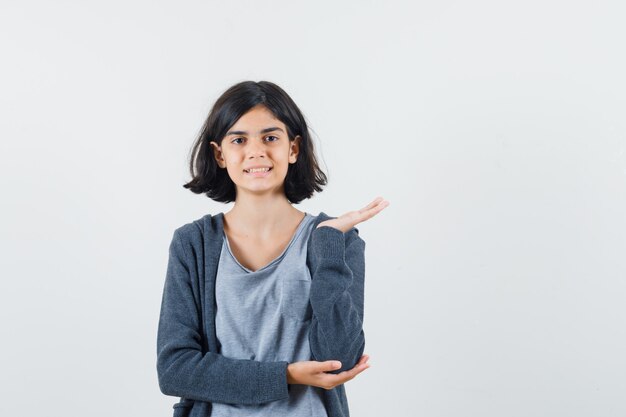 Menina com camiseta e jaqueta fingindo segurar ou apresentar algo e parecendo confiante