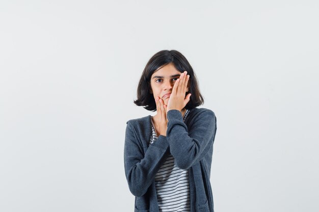 Menina com camiseta, jaqueta segurando a mão na bochecha e parecendo pensativa