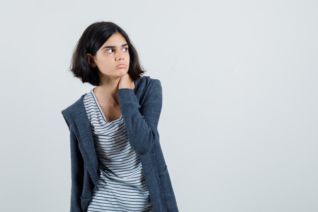 Menina com camiseta, jaqueta segurando a mão no pescoço e parecendo focada,