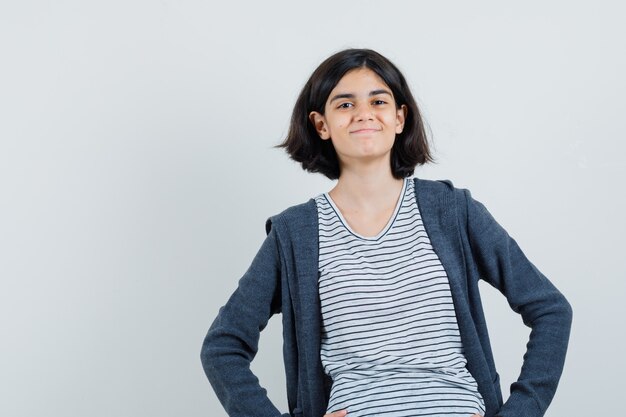 Menina com camiseta, jaqueta segurando as mãos na cintura e parecendo alegre,