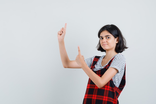 Menina com camiseta, macacão apontando para cima e parecendo atenta,