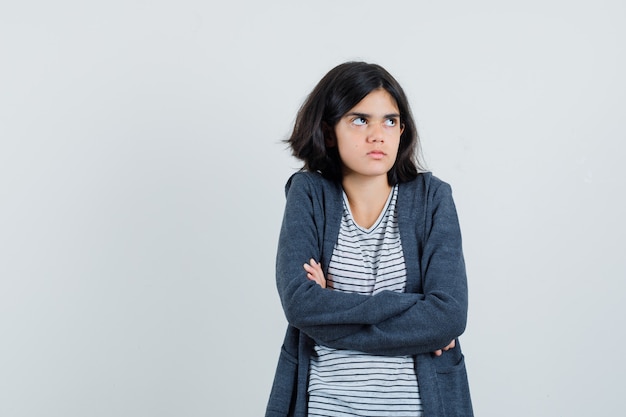 Menina com camiseta, paletó em pé com os braços cruzados e parecendo hesitante,