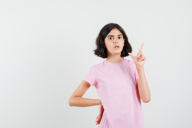 Menina com camiseta rosa apontando para cima e olhando ansiosa, vista frontal.