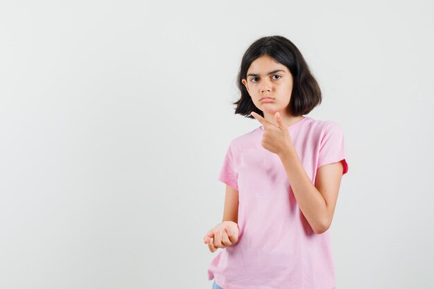 Menina com camiseta rosa apontando para o canto superior esquerdo e olhando séria, vista frontal.