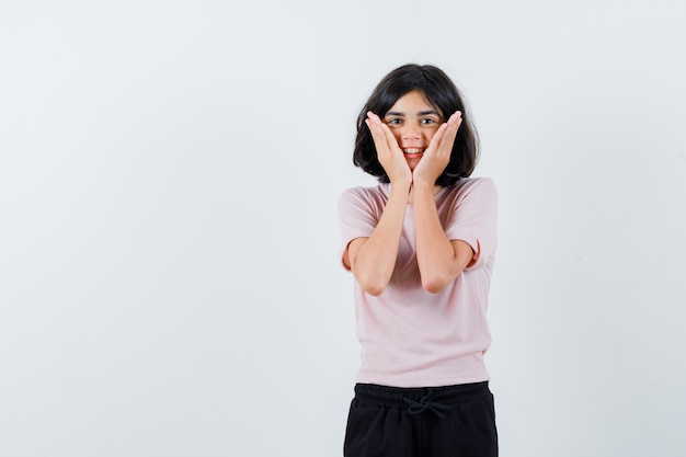 Menina com camiseta rosa e calça preta segurando as mãos no rosto e parecendo uma fofa