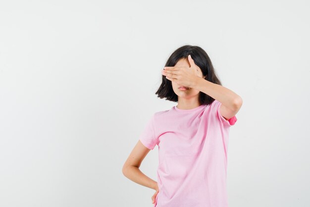 Menina com camiseta rosa, segurando a mão nos olhos e parecendo assustada, vista frontal.