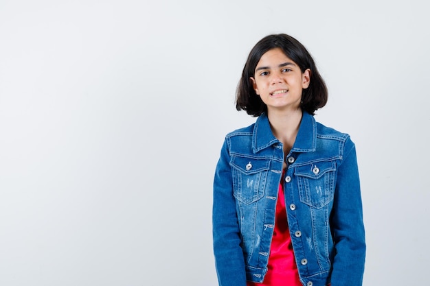 Menina com camiseta vermelha e jaqueta jeans em pé, posando para a câmera e parecendo feliz