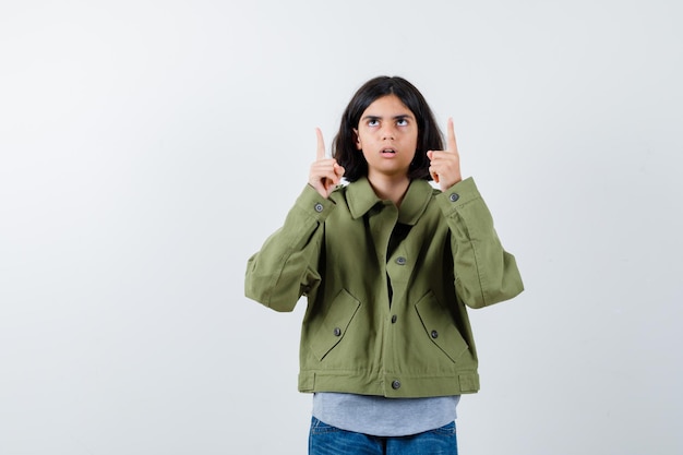 Menina com casaco, camiseta, jeans apontando para cima e olhando com foco, vista frontal.