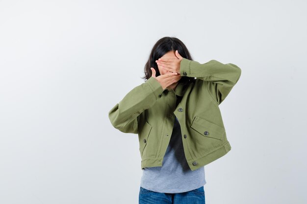 Menina com casaco, camiseta, jeans cobrindo a boca e os olhos com as mãos e olhando bonita, vista frontal.