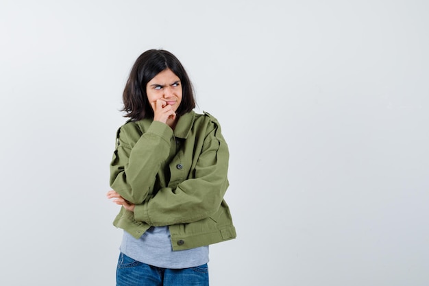 Menina com casaco, camiseta, jeans em pé em pose de pensamento e olhando pensativa, vista frontal.