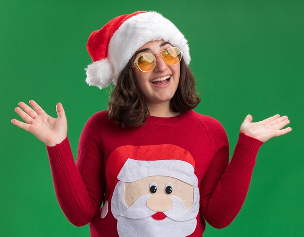 Menina com chapéu de Papai Noel de camisola de natal e óculos olhando para o lado com uma cara feliz levantando as mãos em pé sobre a parede verde