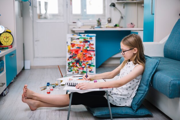 Menina com pintura a lápis na mesa na sala com cores de água no chão