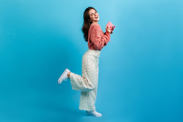 Menina com roupa de luz da moda, posando com câmera rosa na parede azul. a senhora com um sorriso encantador coquete ergueu a perna.