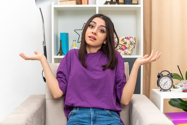 Menina com roupas casuais confusa abrindo os braços para os lados, sentada na cadeira em uma sala iluminada