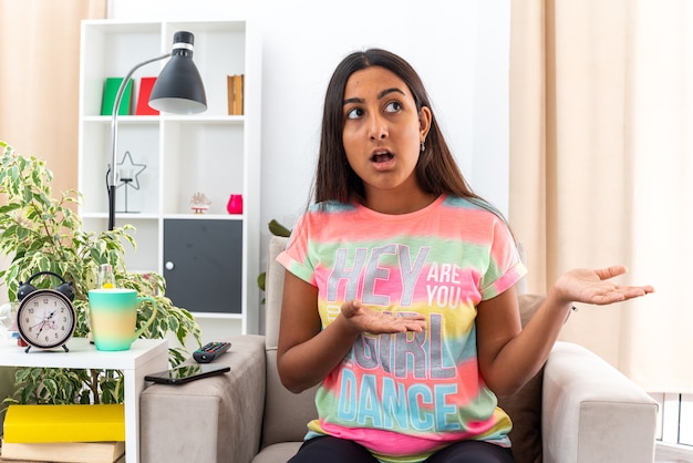 Menina com roupas casuais, olhando de lado, confusa, apresentando-se com o braço das mãos, sentada na cadeira na sala iluminada